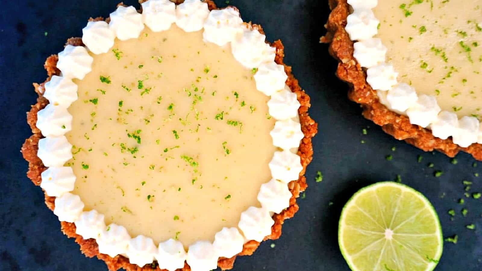 Two mini key lime pies with a graham cracker crust are garnished with a whipped cream border and sprinkled with lime zest. A halved lime is placed beside the pies on a dark surface.