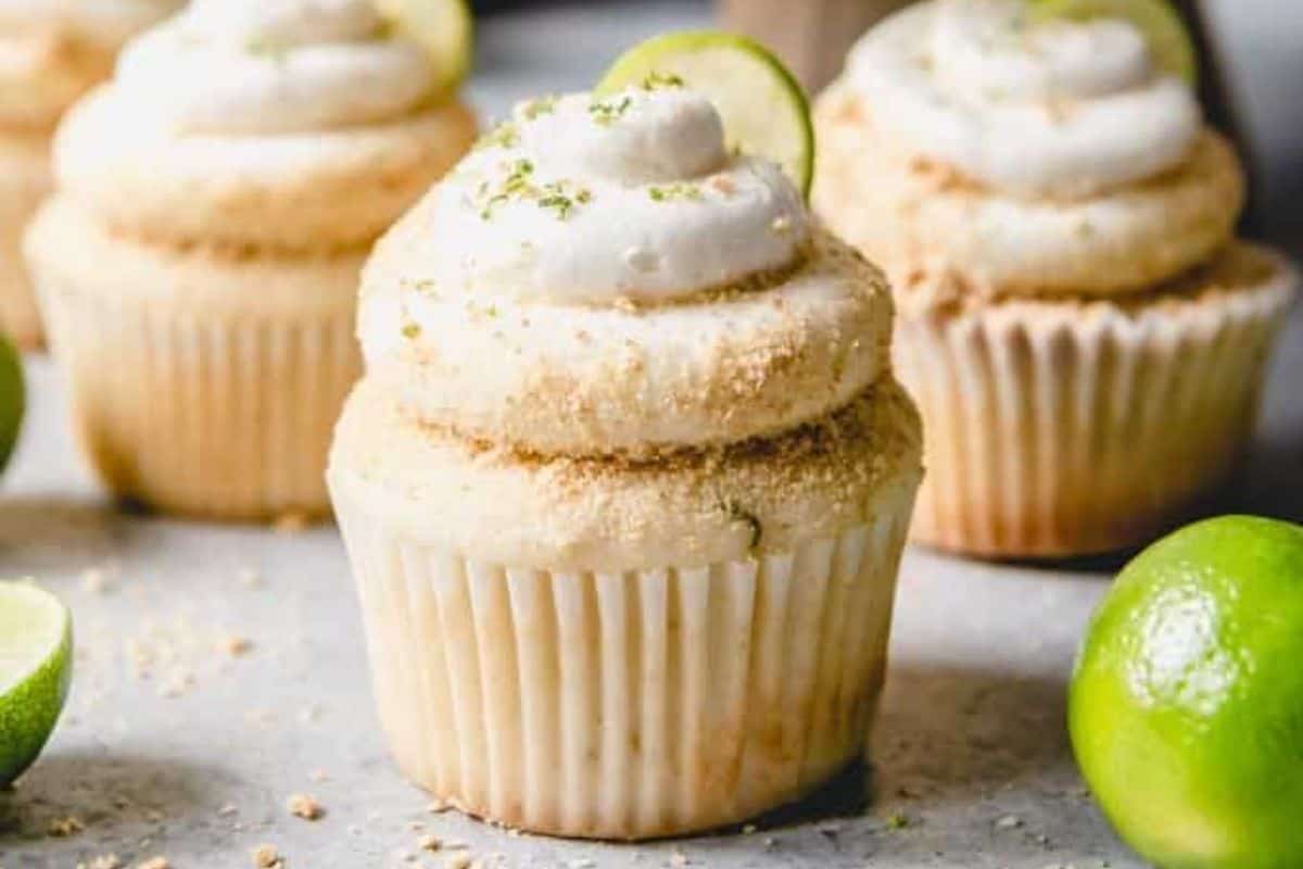 A close-up view of three key lime cupcakes with creamy frosting and sprinkled crumbs on top, set against a light background. Fresh lime slices are used as garnish, adding a touch of vibrant green to the scene. Additional whole limes are scattered around.
