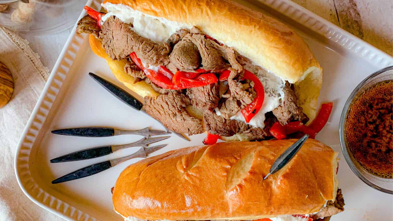 A close-up of two sandwiches filled with sliced roast beef, red bell peppers, and melted cheese, served on a rectangular white plate. Three black-handled knives are placed beside the sandwiches, and a small bowl of au jus dipping sauce is on the side.