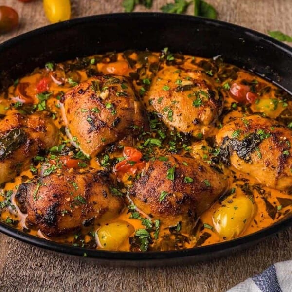 A black roasting pan filled with golden-brown, herb-crusted chicken thighs is garnished with chopped parsley. The dish contains a mixture of colorful cherry tomatoes and roasted vegetables, creating a vibrant and appetizing appearance. The background shows a wooden table with a glimpse of a side dish and some fresh vegetables.