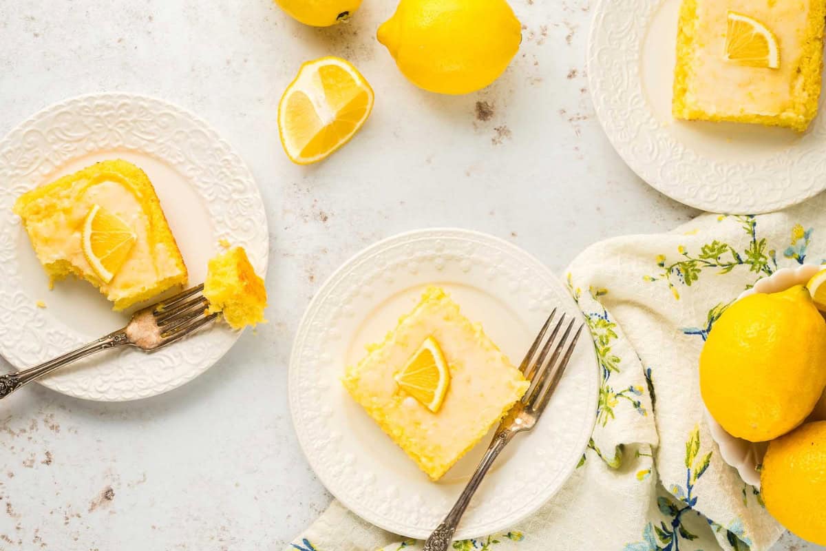 Three plates each hold a slice of lemon cake with a lemon slice garnish. Two forks rest on the side of each plate, with one bite taken from one cake slice. Fresh lemons and a patterned cloth napkin are nearby. The setup is on a light-colored surface.