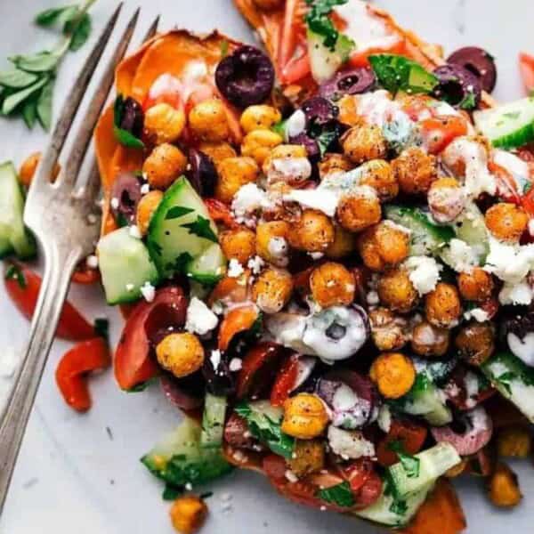 A colorful dish featuring roasted sweet potato halves topped with a vibrant salad of chickpeas, diced cucumbers, tomatoes, olives, feta cheese, and fresh herbs. The plate is garnished with lemon wedges and a fork rests on the side.