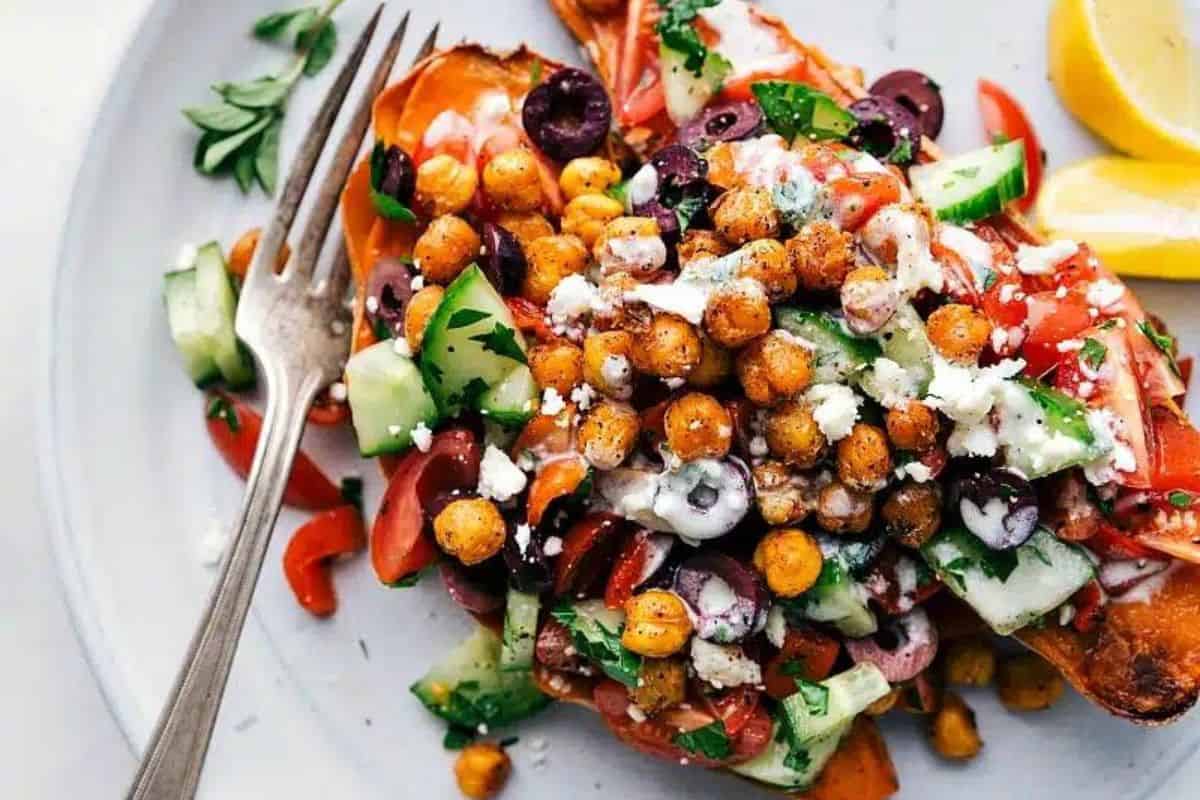 A colorful dish featuring roasted sweet potato halves topped with a vibrant salad of chickpeas, diced cucumbers, tomatoes, olives, feta cheese, and fresh herbs. The plate is garnished with lemon wedges and a fork rests on the side.