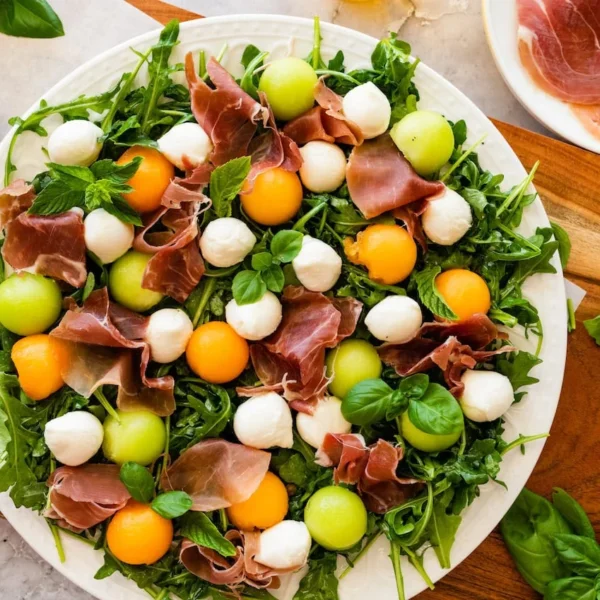 A vibrant salad featuring arugula, slices of prosciutto, balls of mozzarella, and a mix of orange and green melon balls, garnished with fresh basil leaves on a white plate. A small bowl of mozzarella and a plate of prosciutto are in the background.