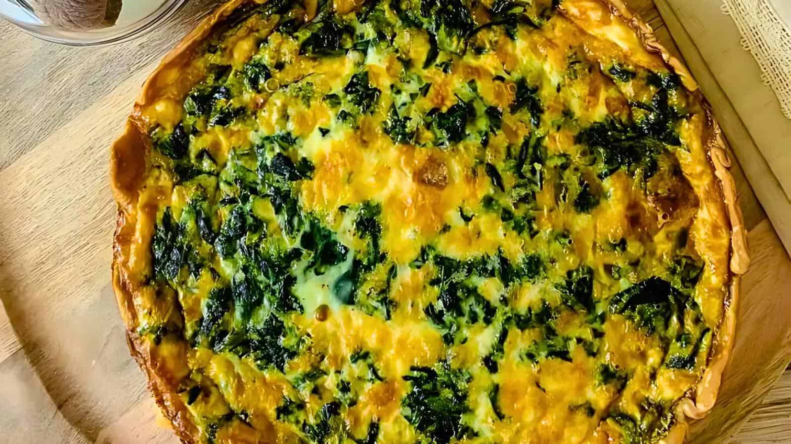 A golden-brown quiche with a flaky crust, filled with spinach and melted cheese, rests on a wooden board.