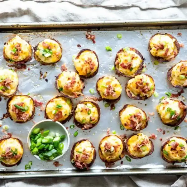 An oven tray filled with numerous bite-sized potato skins, topped with cheese, bacon bits, and green onions. A small bowl of chopped green onions is placed on the tray, and a hand is holding the edge of the tray. The tray is lined with foil.