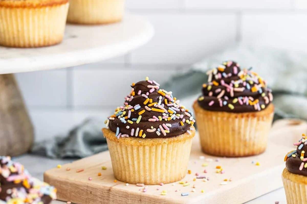 A cupcake with white frosting and multicolored sprinkles, partially wrapped in a paper liner, on a mesh surface.