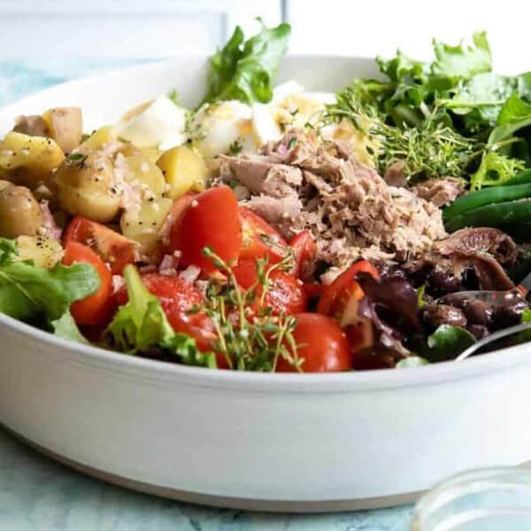 A colorful salad bowl featuring fresh lettuce, boiled potatoes, tomatoes, green beans, tuna, and olives, garnished with herbs. Chopsticks are resting on the edge of the bowl, with a light blue background.