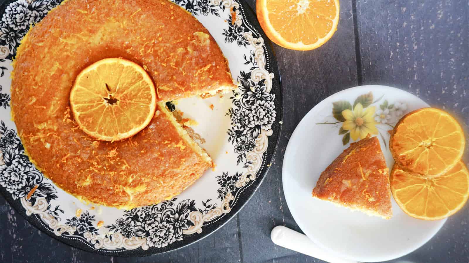 A slice of cake on a plate with a larger plate with more cake decorated with a slice of orange.