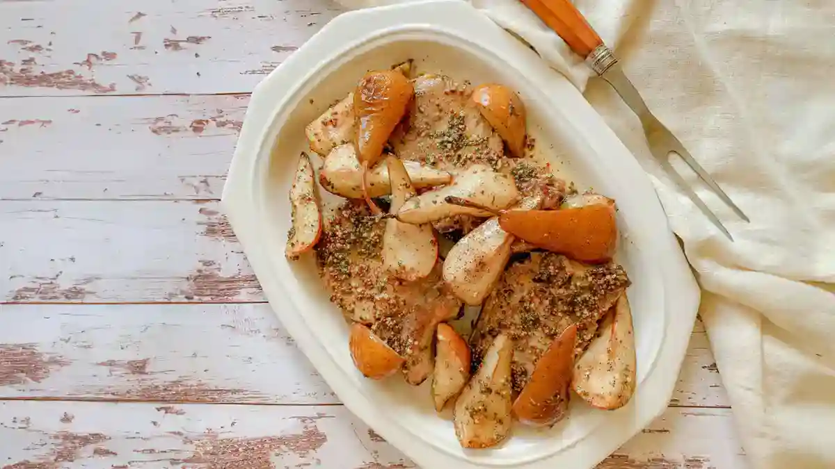 A white oval plate with baked chicken and pears, seasoned with herbs, on a rustic wooden table beside a carving fork and a beige napkin.