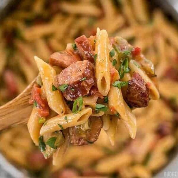 A wooden spoon holds up a serving of penne pasta mixed with chunks of sausage, diced tomatoes, green bell peppers, and herbs. The background shows a pan filled with the same pasta dish, slightly out of focus.