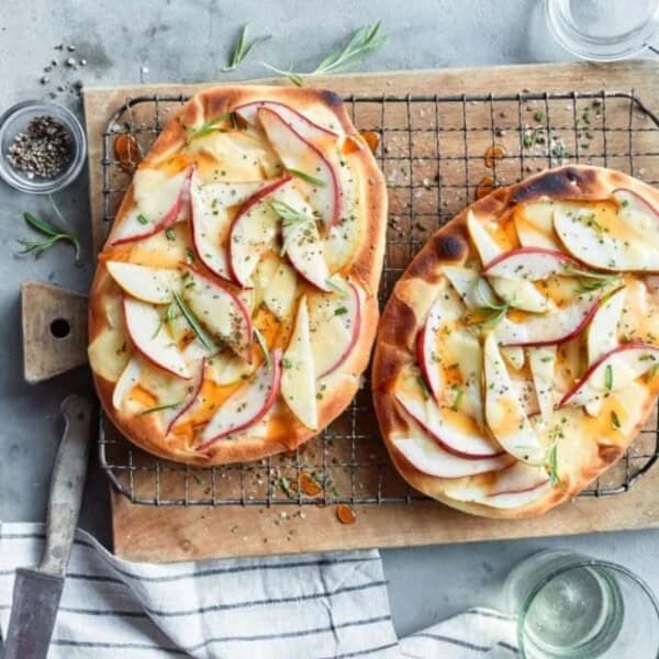 Two flatbreads topped with sliced apples, herbs, and melted cheese are placed on a wire rack over a wooden cutting board. Surrounding the flatbreads are a knife, salt, pepper, a small jar of honey, and empty glass jars. A striped cloth lies partially in view.