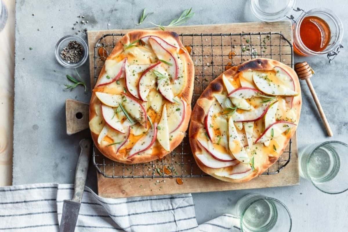 Two flatbreads topped with sliced apples, herbs, and melted cheese are placed on a wire rack over a wooden cutting board. Surrounding the flatbreads are a knife, salt, pepper, a small jar of honey, and empty glass jars. A striped cloth lies partially in view.