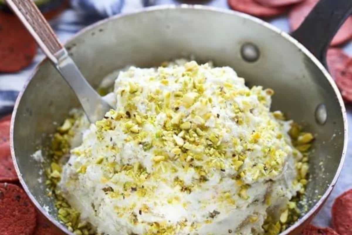 A pot filled with creamy dessert topped with chopped pistachios. A spatula is resting in the pot, and red round biscuits surround the pot.