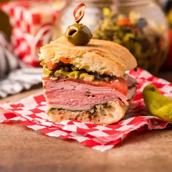 A thick muffuletta sandwich with cold cuts, cheese, and olive salad is topped with a green olive on checkered paper by a pepper.