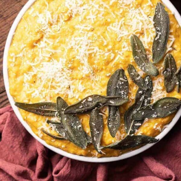A bowl of risotto with grated cheese and sage leaves on a wooden table next to a pink cloth and a pinecone.
