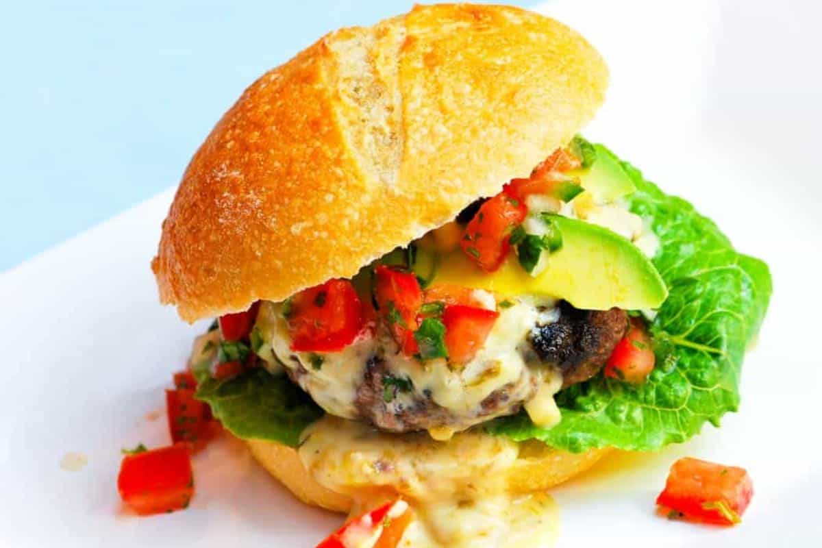 A gourmet hamburger with a sesame seed bun, topped with fresh tomatoes, avocado slices, lettuce, and drizzled with a creamy sauce. The burger has combined textures and colors, showcasing a mix of juicy vegetables and crispy lettuce.