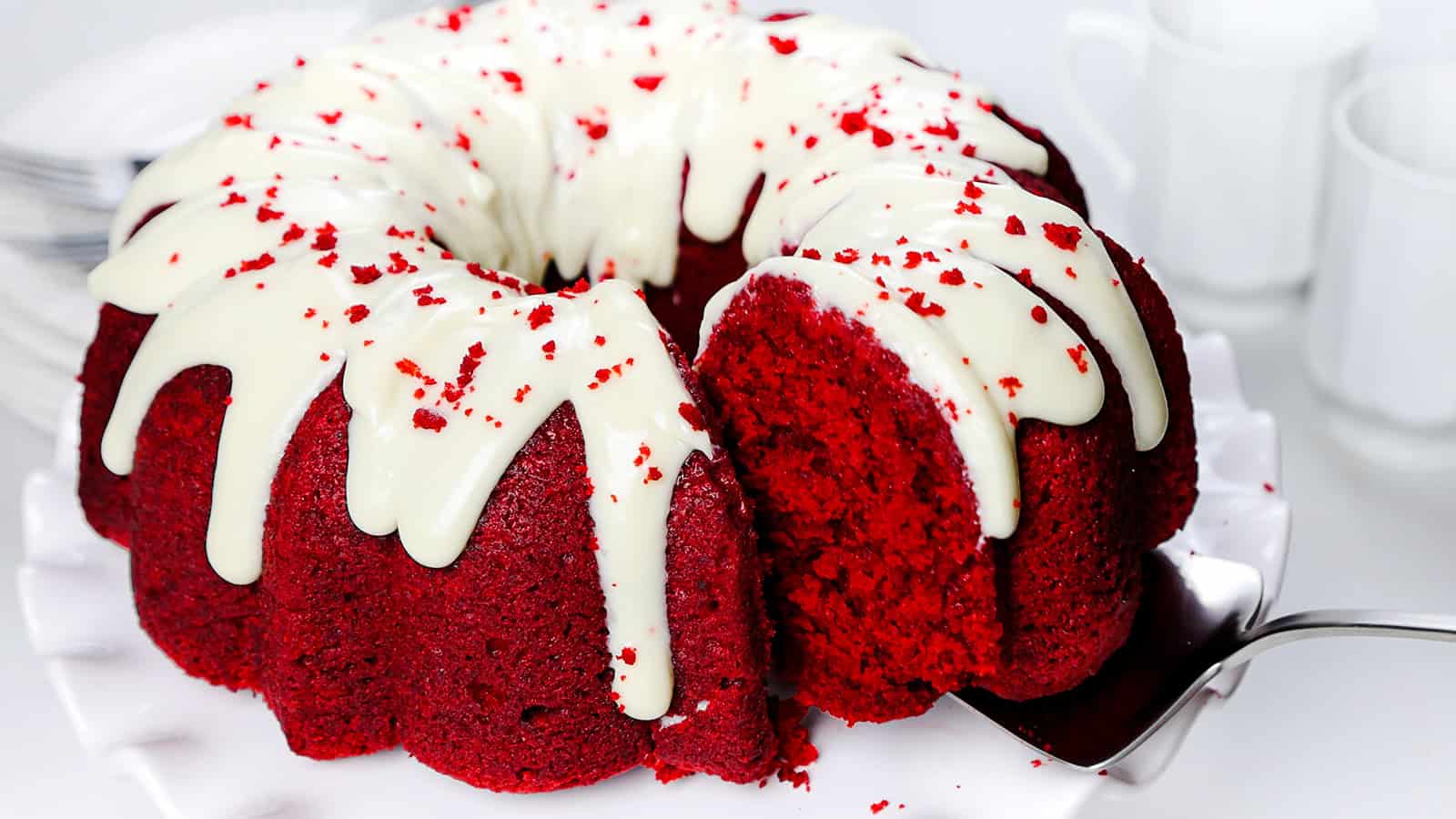 A close-up of a red velvet Bundt cake topped with white cream cheese frosting, partially sliced with a piece being lifted out on a serving utensil. The cake sits on a white plate, and the background features blurred white cups.
