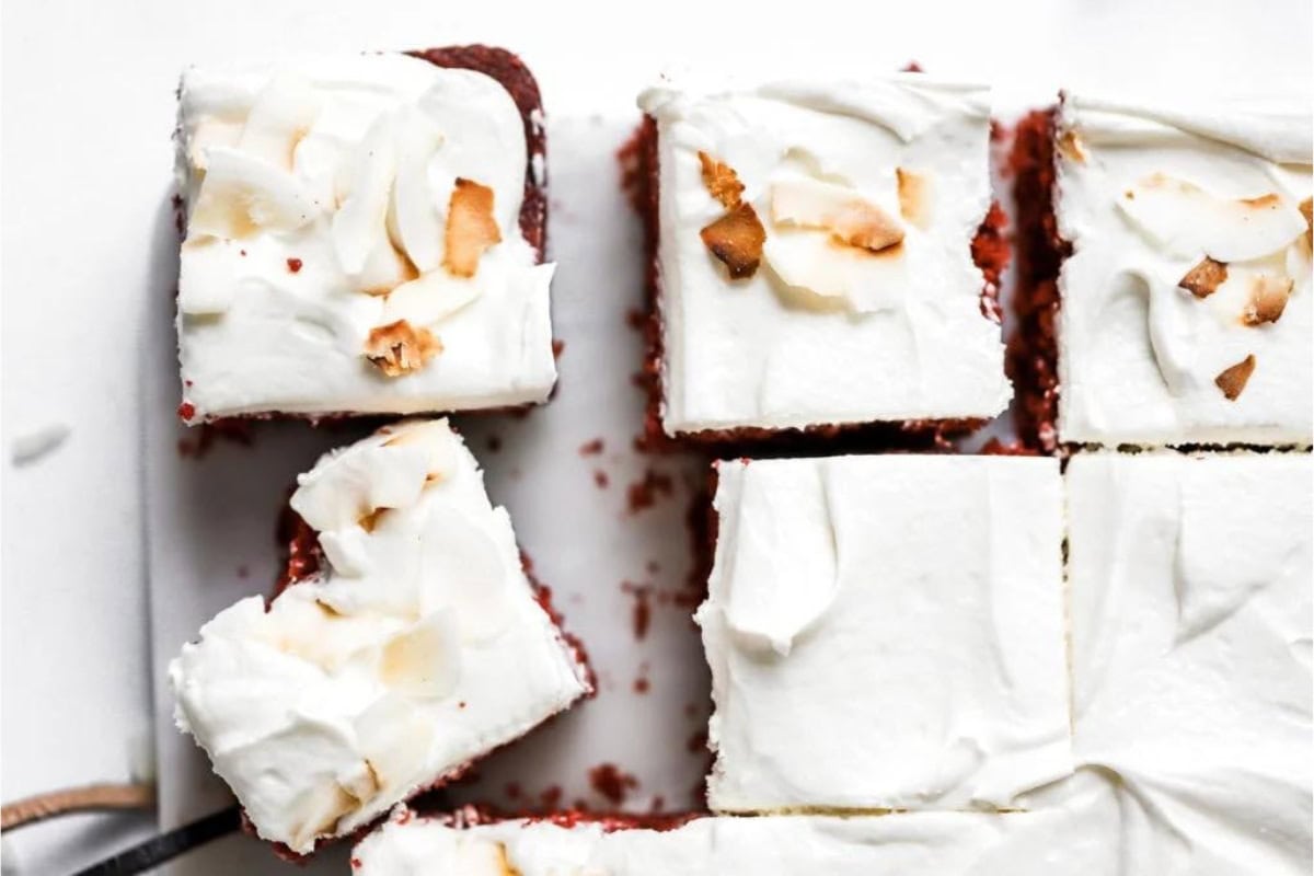 A close-up view of six square pieces of red velvet cake topped with white frosting and coconut flakes. One piece has a bite taken out of it, revealing the red cake inside. The pieces are arranged on a white surface.