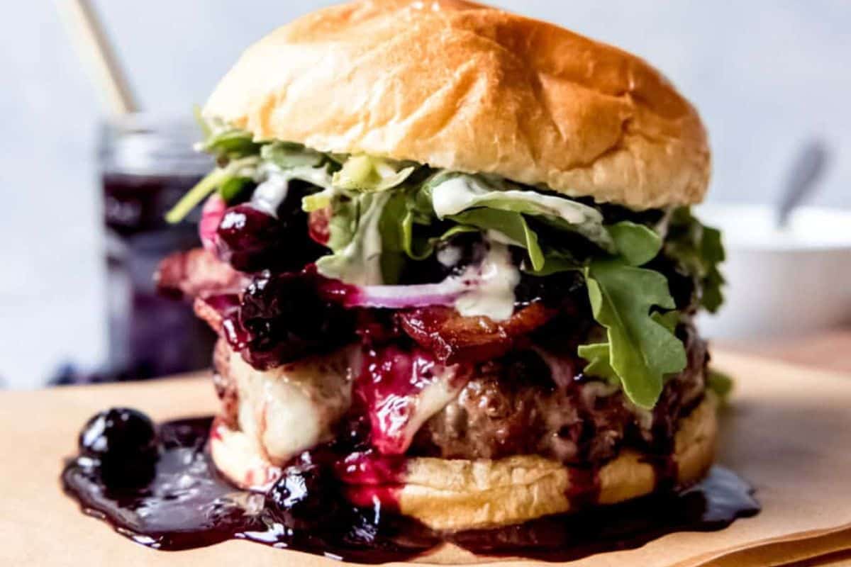A juicy burger topped with melted cheese, mixed greens, slices of red onion, and a generous amount of blueberry sauce on a toasted bun. The burger is presented on a piece of parchment paper, with a jar of blueberry sauce and a blurred bowl in the background.
