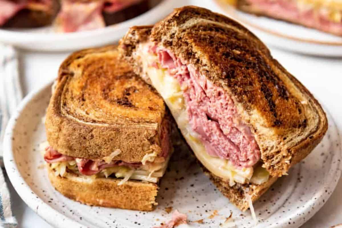 A close-up of a Reuben sandwich on a speckled ceramic plate. The sandwich is made with rye bread and filled with corned beef, sauerkraut, and melted cheese, neatly cut in half and stacked to show the layers. Another sandwich is visible in the background.
