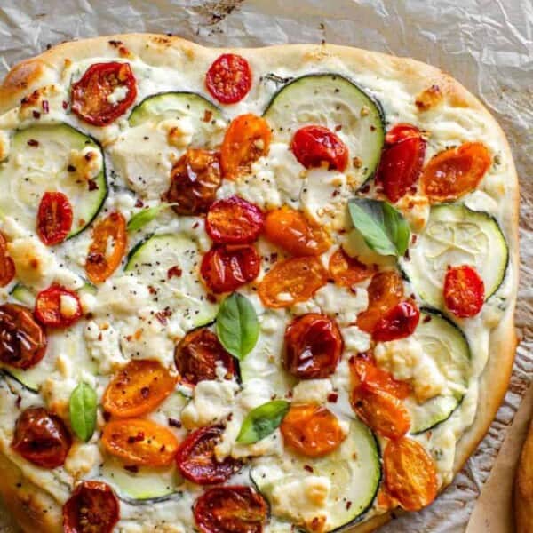 A rectangular flatbread on parchment paper, topped with sliced zucchini, halved cherry tomatoes, crumbled cheese, and fresh basil.