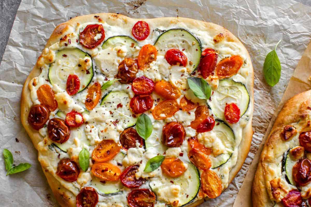 A freshly baked flatbread pizza topped with sliced zucchini, halved cherry tomatoes, and crumbled feta cheese. The pizza rests on parchment paper, garnished with a few fresh basil leaves, and there is a portion of another pizza visible on the side.