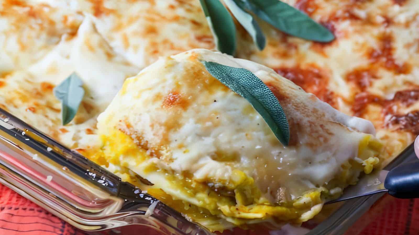 A slice of cheesy, golden-brown lasagna topped with fresh sage is being lifted from a glass baking dish. The lasagna layers are visible, showcasing creamy sauce and pasta. The dish is presented on a red cloth, enhancing the visual appeal of the warm, baked meal.