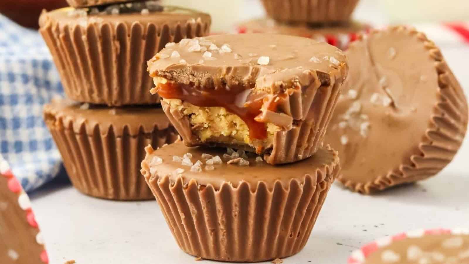 Close-up of stacked chocolate cups with one cut open, revealing caramel and nut filling. A sprinkle of sea salt is visible on top. A checkered napkin is in the background.