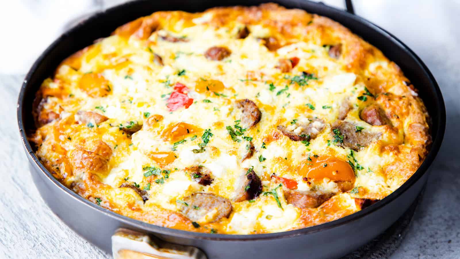 A close-up of a freshly cooked frittata in a black skillet. The frittata is golden brown on the edges and features ingredients like sausage, cherry tomatoes, herbs, and melted cheese. The handle of the skillet is visible in the foreground.