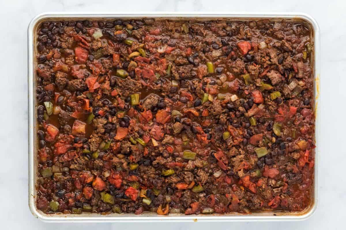 A sheet pan with cooked ground beef, black beans, chopped tomatoes, and green bell peppers mixed together in sauce.