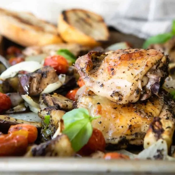 A tray of roasted chicken pieces with tomatoes, onions, and eggplant, garnished with fresh basil leaves. Grilled bread is visible in the background.