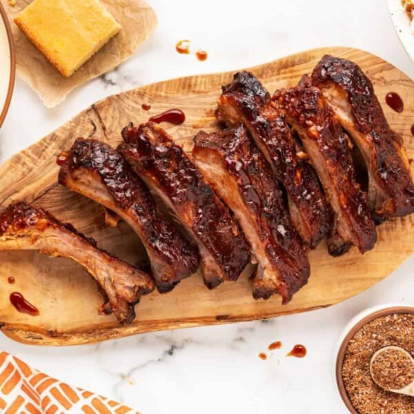 Barbecued ribs on a wooden board with cornbread, coleslaw, spice rub, forks, and an orange patterned napkin nearby.