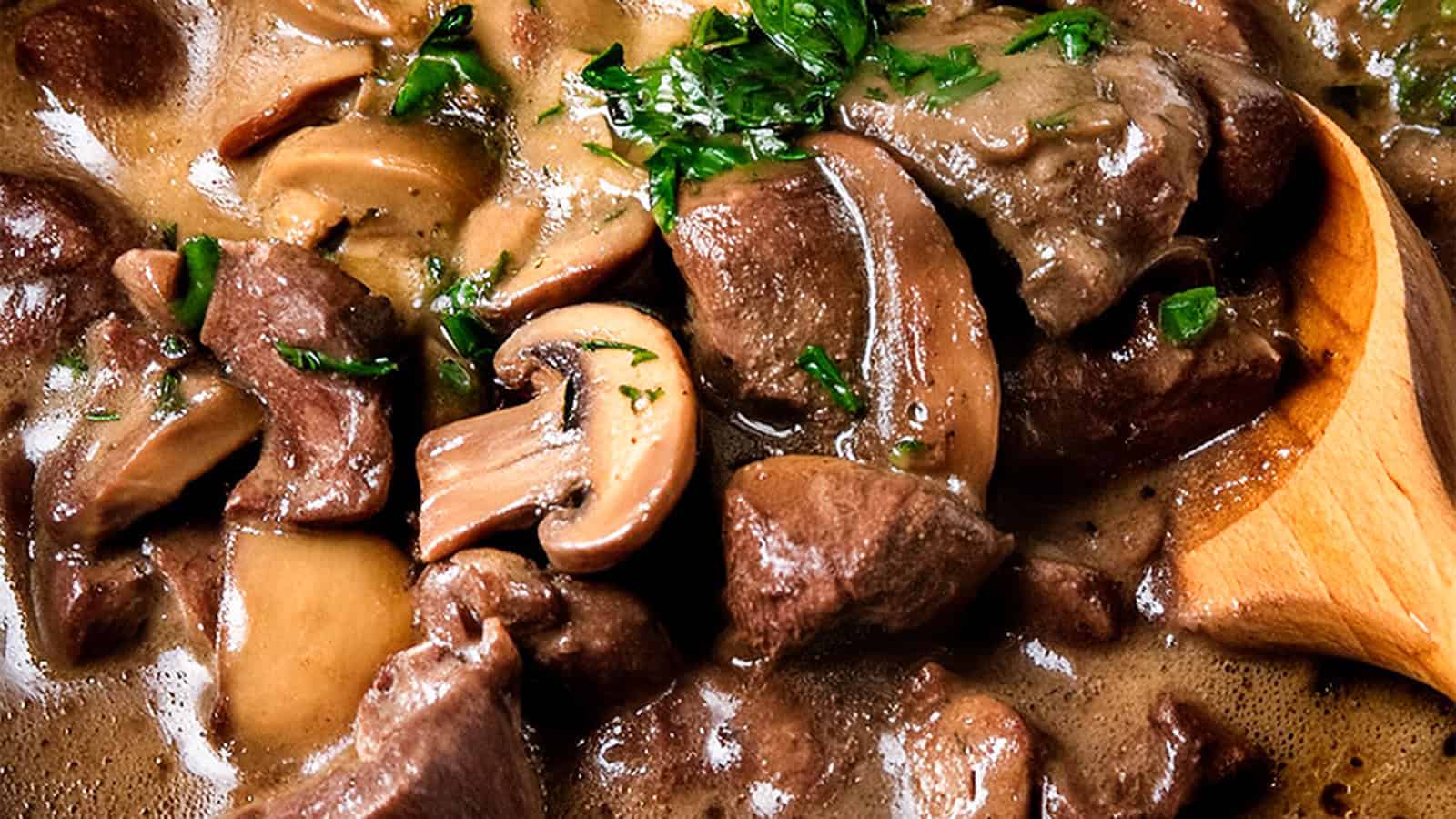 A savory dish of beef tips and mushrooms is shown in a creamy, dark sauce. Freshly chopped parsley is sprinkled on top for garnish, and a wooden spoon is partially visible in the bottom right corner. The dish appears hearty and comforting.