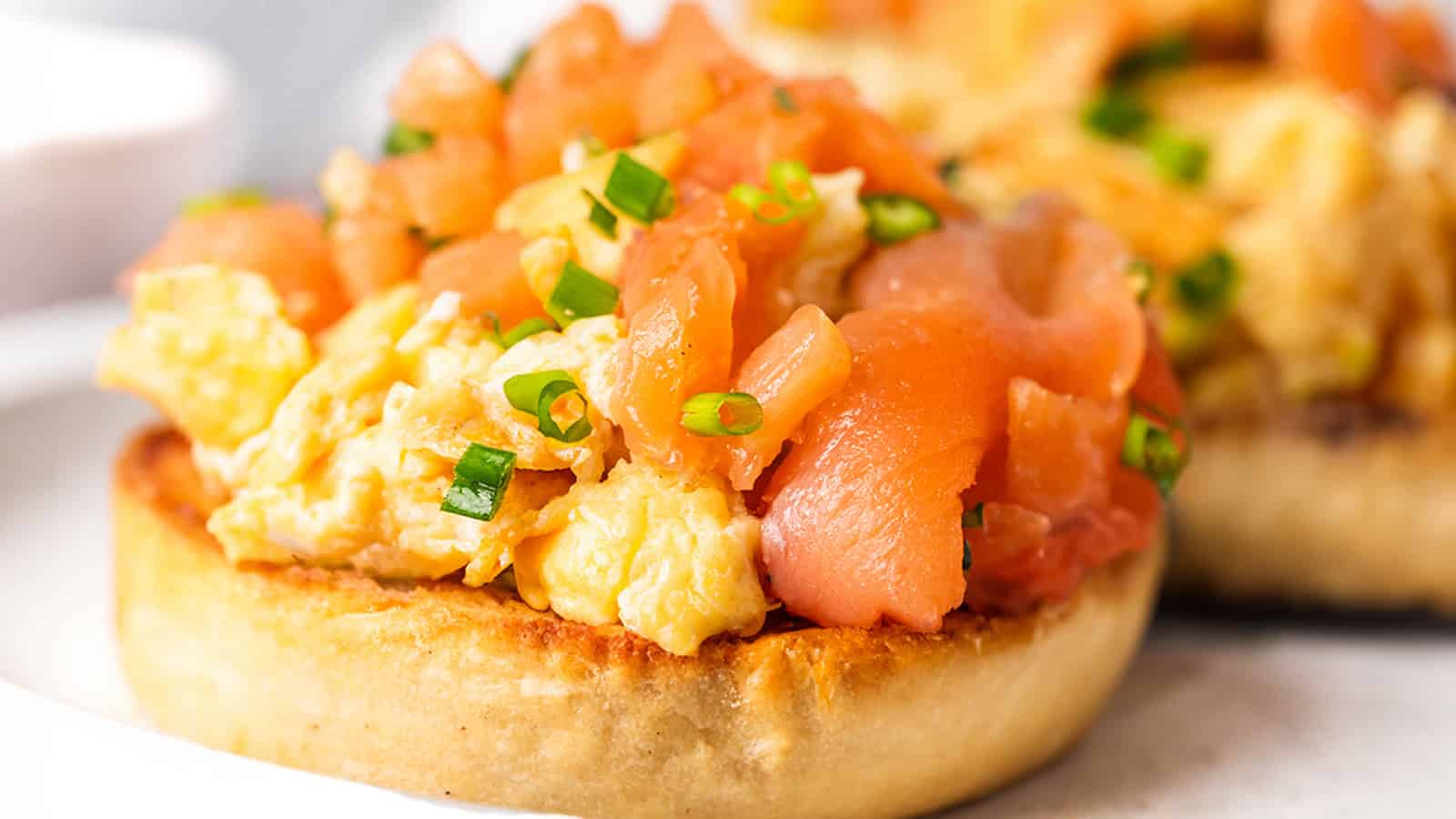 Close-up of a toasted bagel half topped with scrambled eggs, slices of smoked salmon, and garnished with chopped green onions. The background is blurred, emphasizing the focus on the bagel.