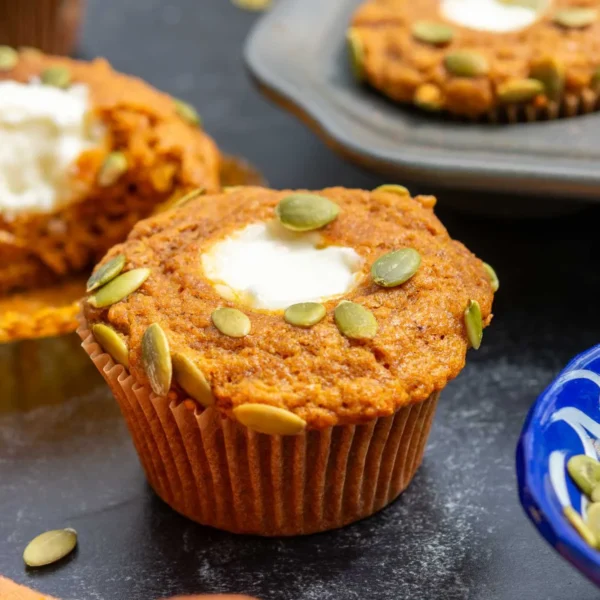 A pumpkin muffin with a cream cheese center and pumpkin seeds on top sits on a dark surface, with more muffins and a bowl of seeds nearby.