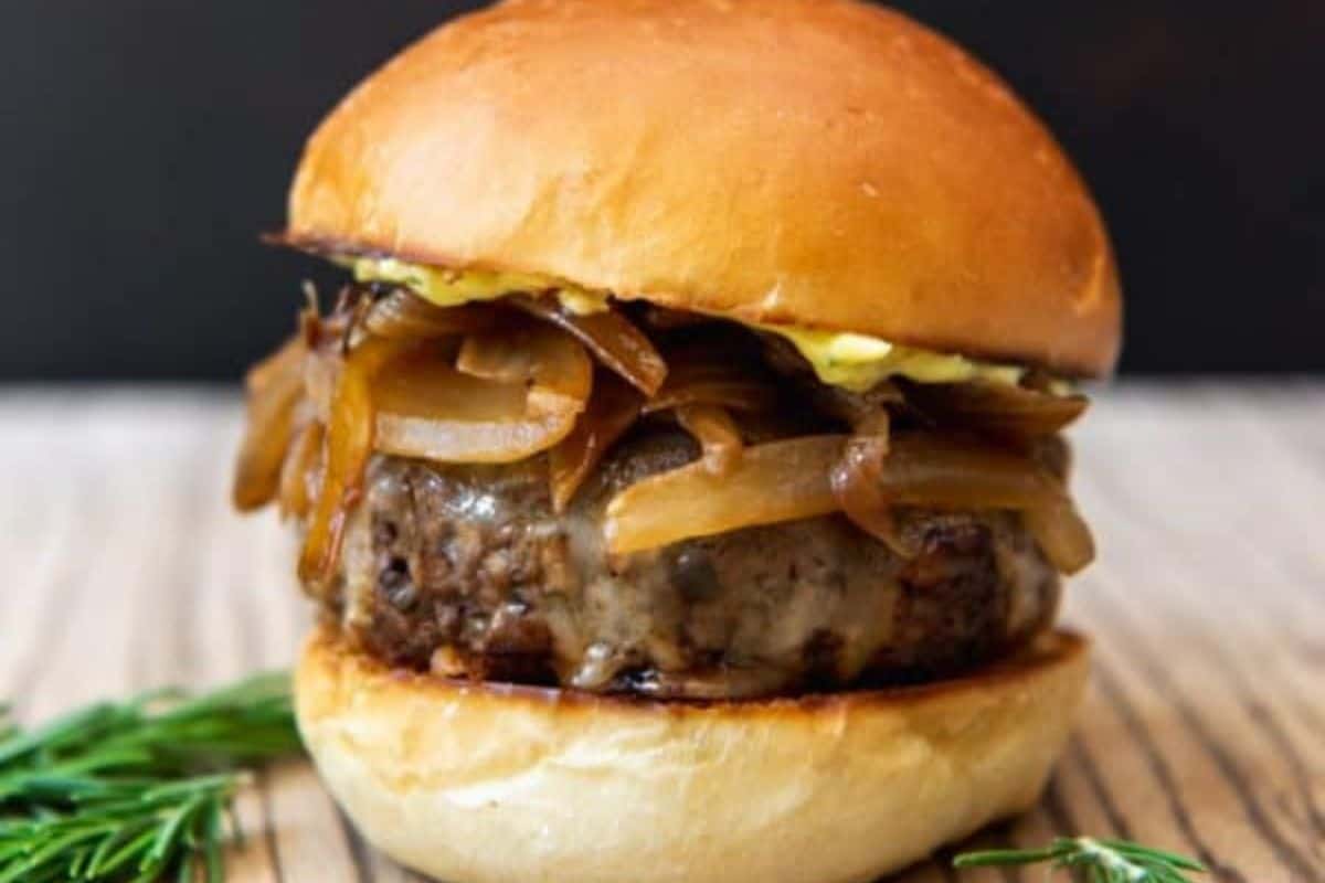 A close-up of a gourmet burger on a wooden surface. The burger features a juicy beef patty topped with caramelized onions and drizzled with a light sauce, all encased in a glossy, golden-brown bun. Fresh rosemary sprigs are seen in the foreground.