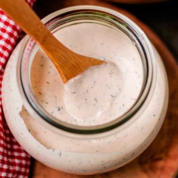 A glass jar filled with creamy ranch dressing mixed with herbs, a wooden spoon inside, and a red and white checkered cloth beside it.