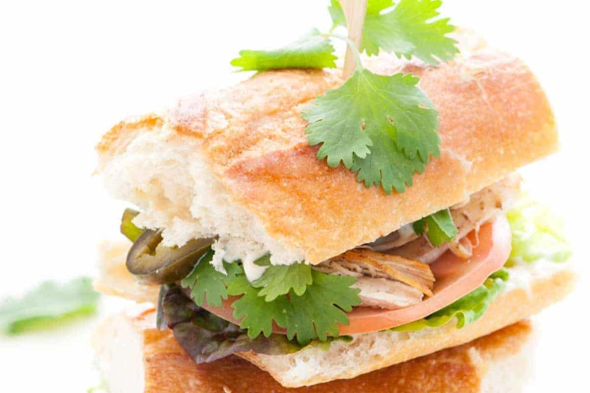 A close-up of a sandwich made with a crusty baguette, filled with shredded meat, tomato slices, leafy greens, jalapeño slices, and fresh cilantro sprigs on top. The sandwich is cut in half, with one half resting on the other.