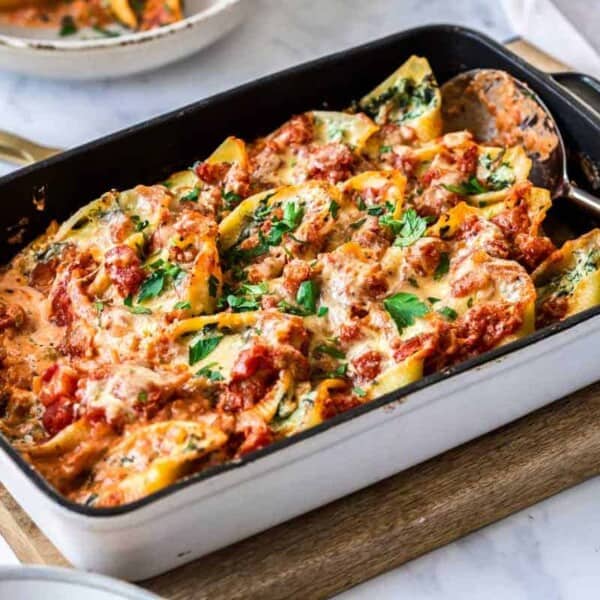 A white rectangular baking dish filled with stuffed manicotti pasta topped with melted cheese and marinara sauce, garnished with fresh parsley. The dish rests on a wooden cutting board, with a serving spoon on the side.