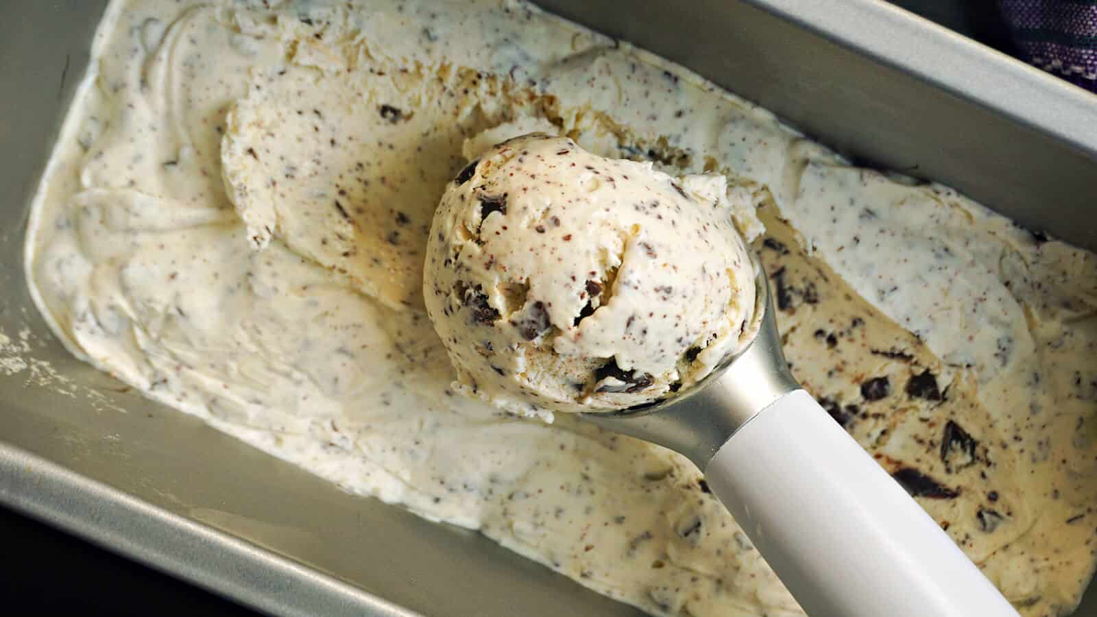 A scoop of cookies and cream ice cream is being lifted from a rectangular metal container with a white-handled ice cream scoop. The ice cream is creamy with visible chunks of chocolate cookies.