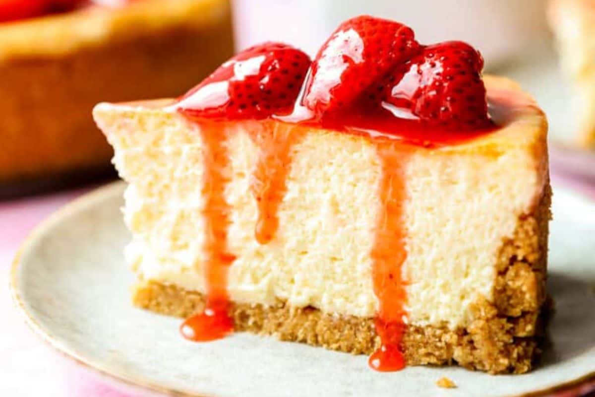 A close-up of a slice of cheesecake on a plate, topped with a generous amount of bright red strawberry sauce and fresh strawberry slices. The cheesecake has a thick, creamy layer and a crumbly, golden-brown graham cracker crust.