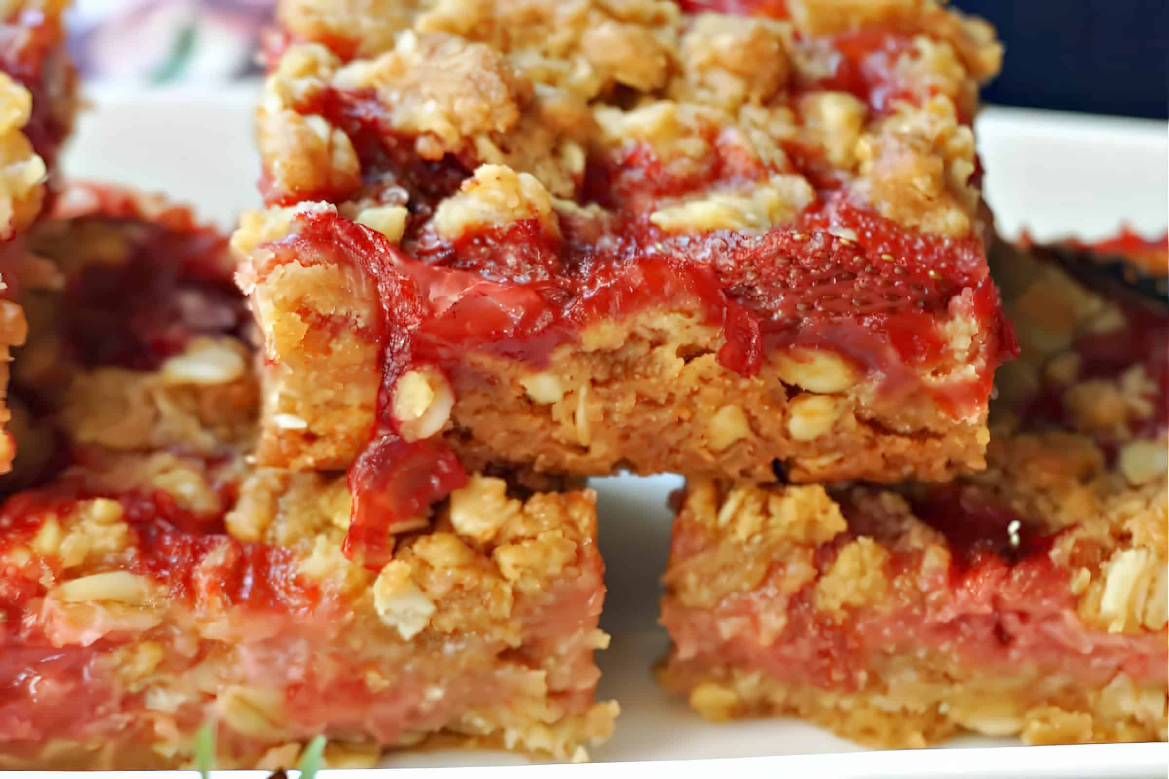 Close-up of several layered strawberry oatmeal bars. The bars have a crumbly oat and brown sugar crust with a bright red strawberry filling that appears moist and juicy. The texture of the oats and the vibrant color of the strawberries are prominent.