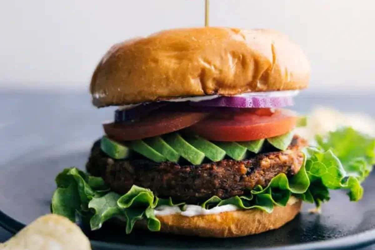 A close-up of a veggie burger on a plate. The burger is stacked with layers of lettuce, avocado slices, tomato, and red onion, all sandwiched between a shiny, golden-brown bun. A toothpick is inserted through the center to hold the ingredients together.