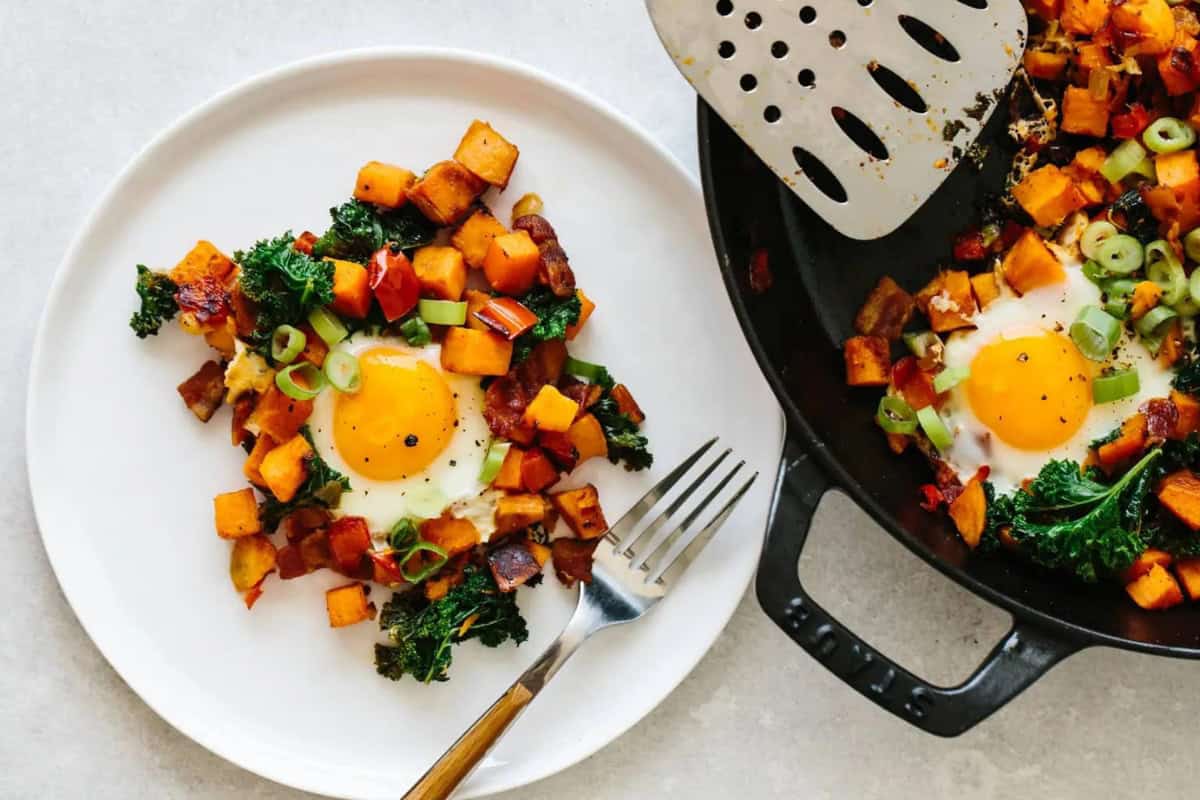 A plate with a serving of sweet potato hash featuring a sunny-side-up egg, sautéed kale, and diced bell peppers. A skillet with more hash, an egg, and a spatula lies next to the plate. A fork rests on the plate.