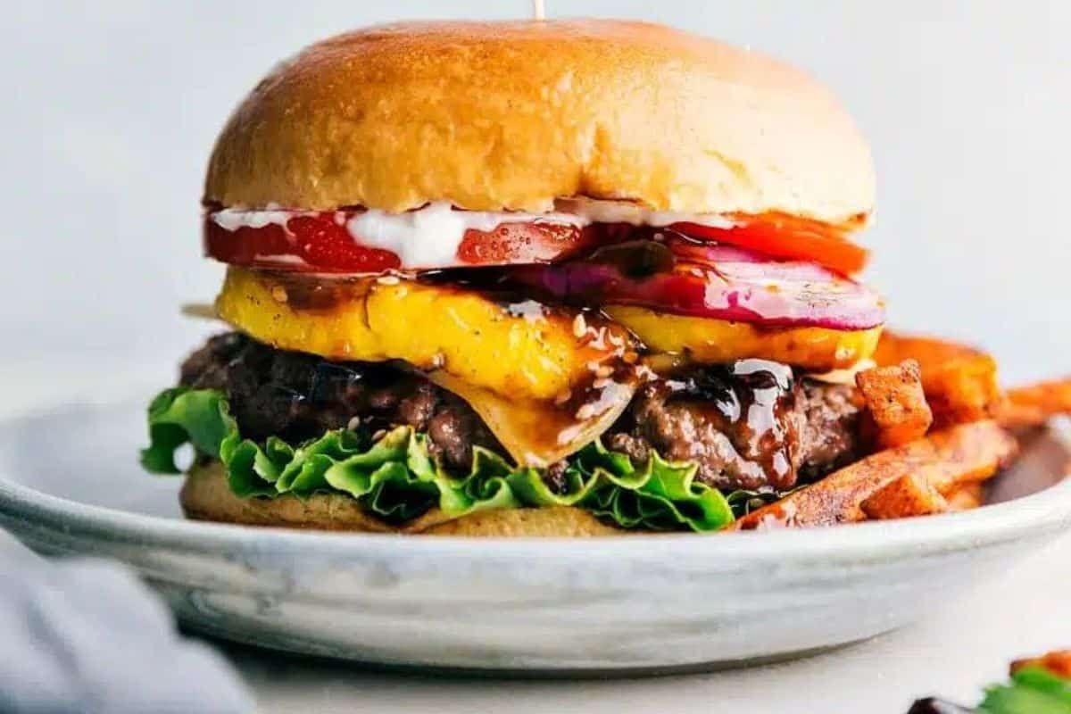 A close-up of a burger with a beef patty, lettuce, melted cheese, grilled pineapple, red onions, tomato slices, and sauce, all inside a shiny toasted bun. The burger is served on a plate with a few fries visible on the side.