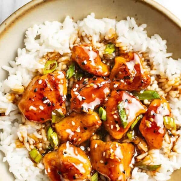 A bowl filled with white rice topped with glazed chunks of chicken, sprinkled with sesame seeds and chopped green onions.