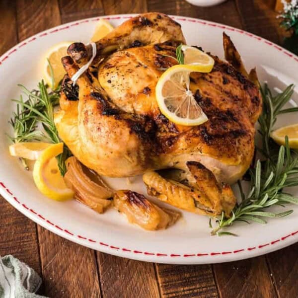Roast chicken with lemon slices and rosemary on a white oval platter, surrounded by roasted onions and lemon wedges.