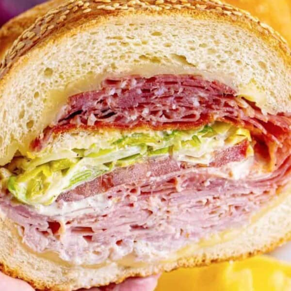 A close-up of a thick sandwich held in hand, showcasing layers of sliced salami, capicola, and ham, with shredded lettuce, tomato slices, and a spread of mayonnaise, all encased in a sesame seed bun.
