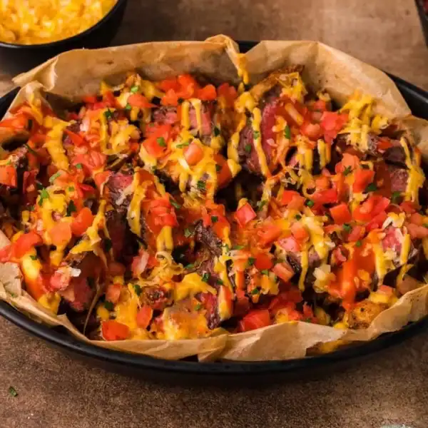 A black dish with parchment paper holds steak nachos topped with melted cheese, diced tomatoes, and herbs. Bowls sit behind it.